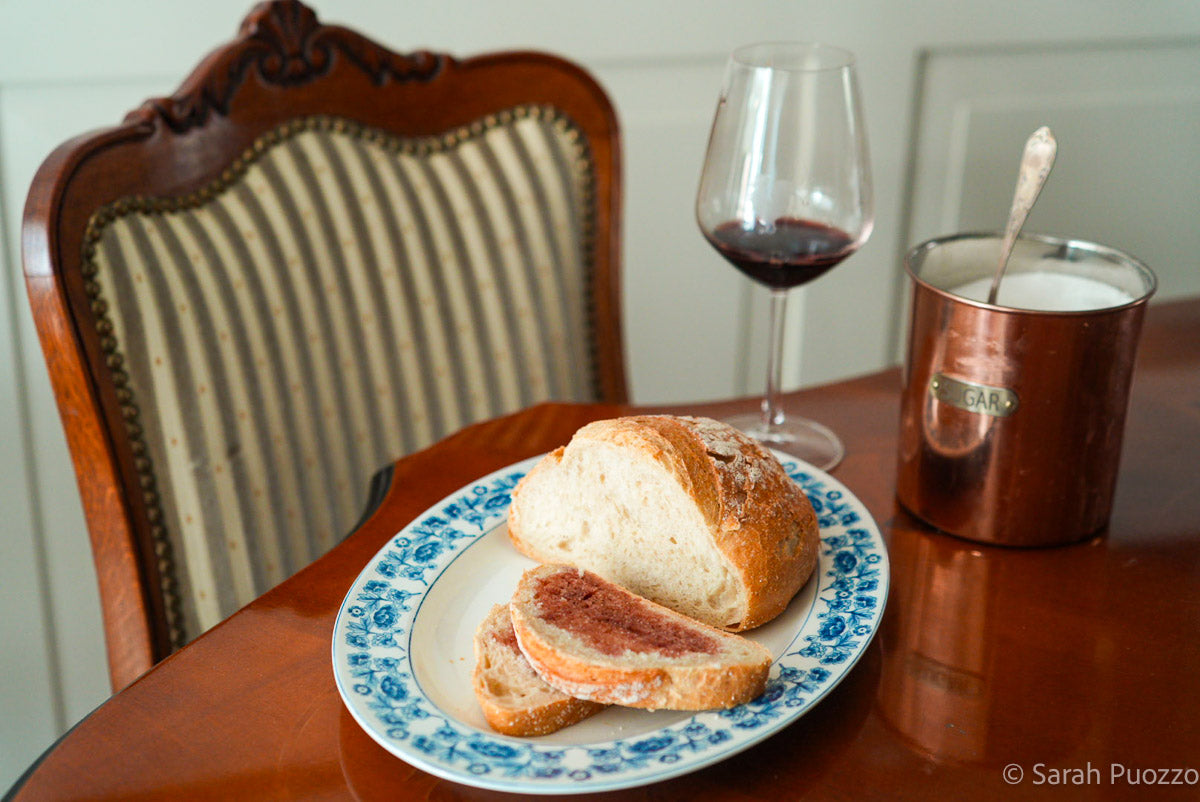 Een tafel gedekt met een schaal brood, een boterham met rode wijn en suiker erop, met op de achtergrond een pot met suiker en een glas rode wijn.